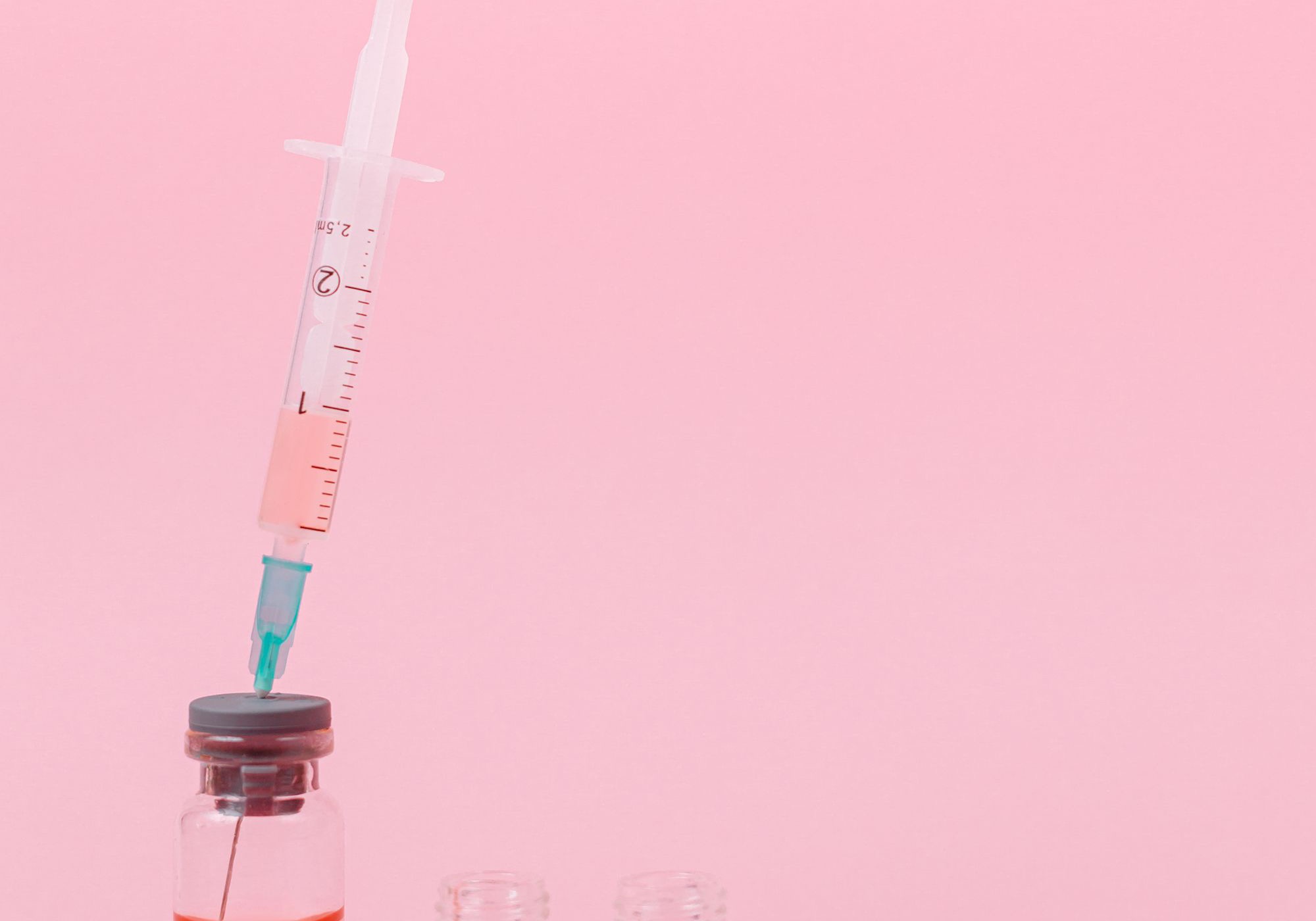 A graduated syringe drawing pink liquid from a small rubber-capped glass vial, with two empty vials on a plain pink surface, evoking peptide reconstitution