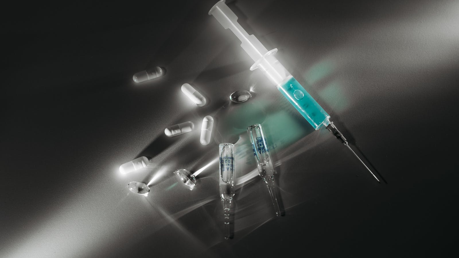 A syringe of teal liquid beside scattered translucent capsules and glass ampoules on a dark reflective surface