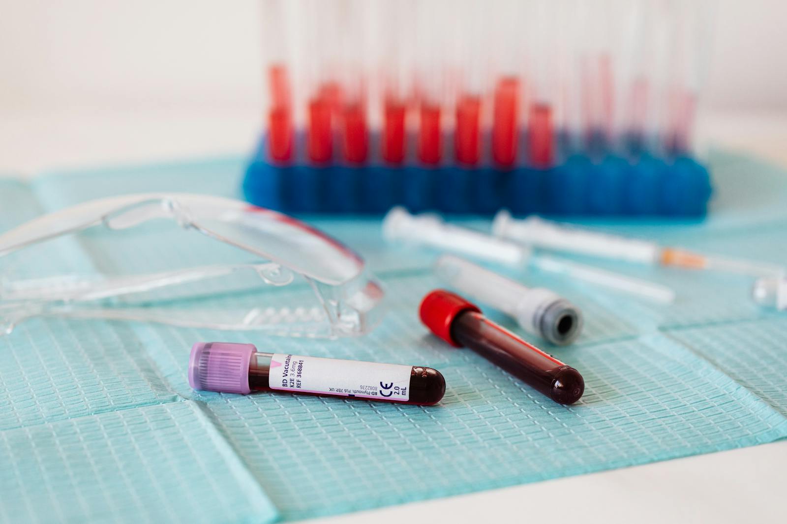 Filled blood collection tubes on a clinical drape beside safety glasses and syringes, with a rack of sample tubes behind
