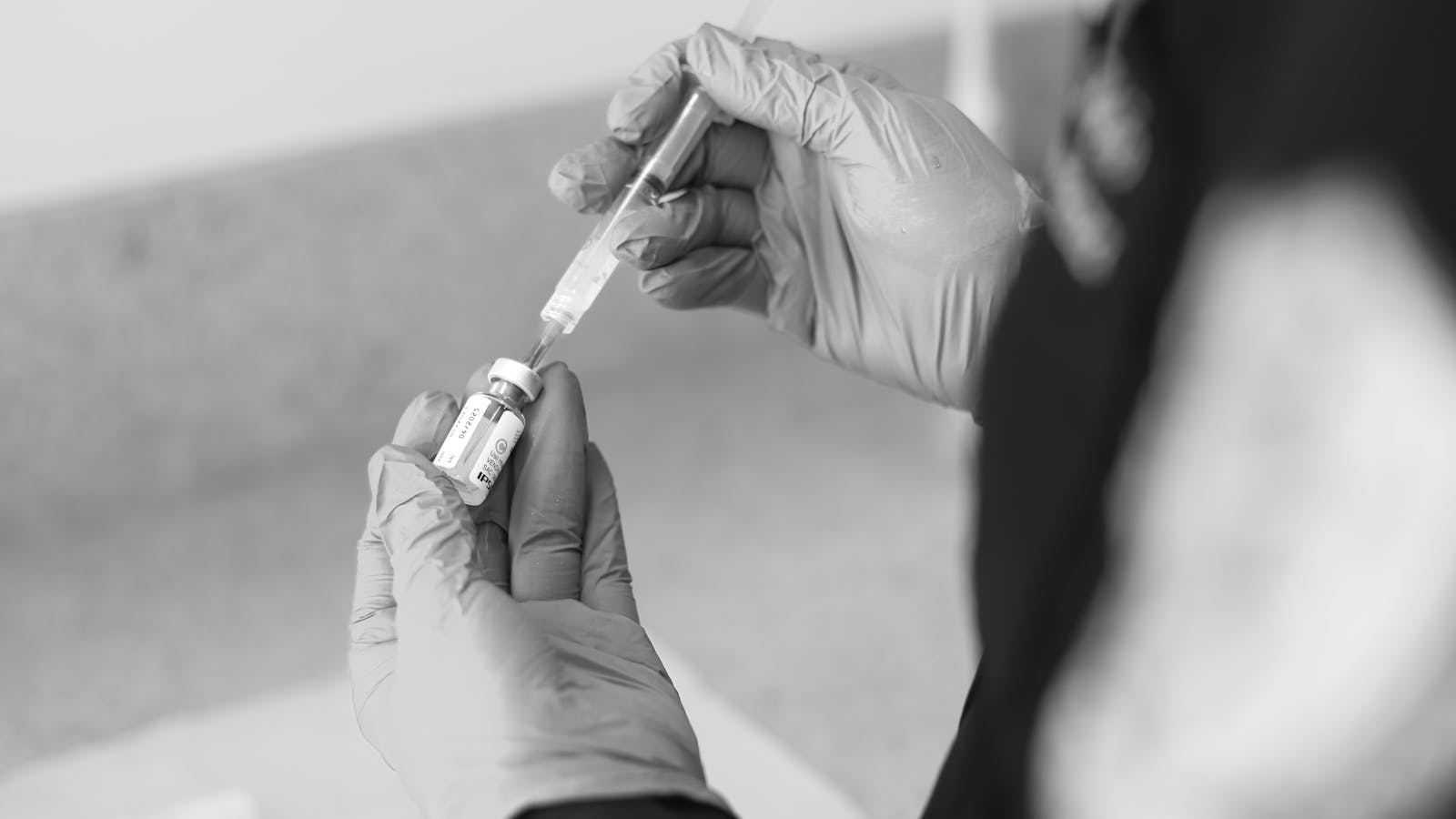 Black-and-white close-up of gloved hands inserting a syringe into a small medication vial