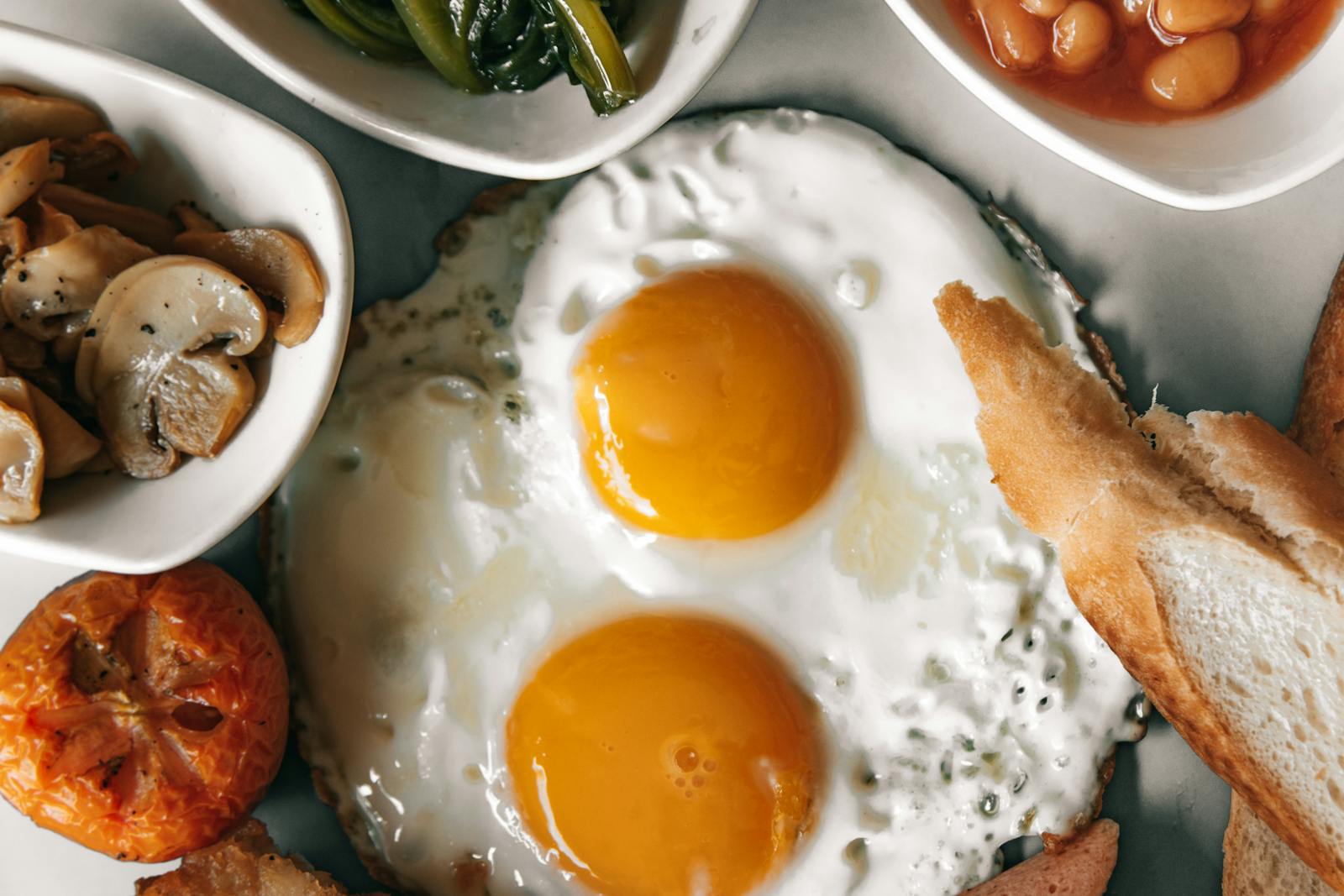 Top-down view of a cooked breakfast plate with two sunny-side-up eggs, mushrooms, beans, greens, tomato, and bread