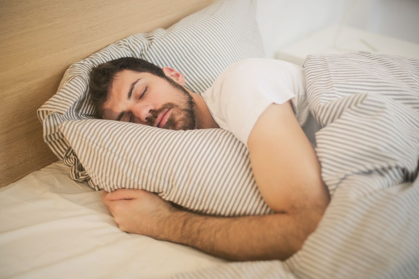 A man sleeping peacefully on striped bedding in soft light, evoking the deep-sleep growth-hormone pulse that ipamorelin is dosed pre-bed to align with