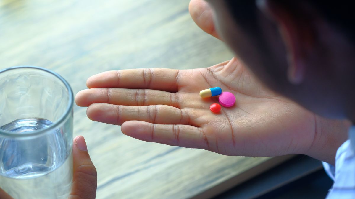 A hand holding a pill beside a clear glass of water on a table