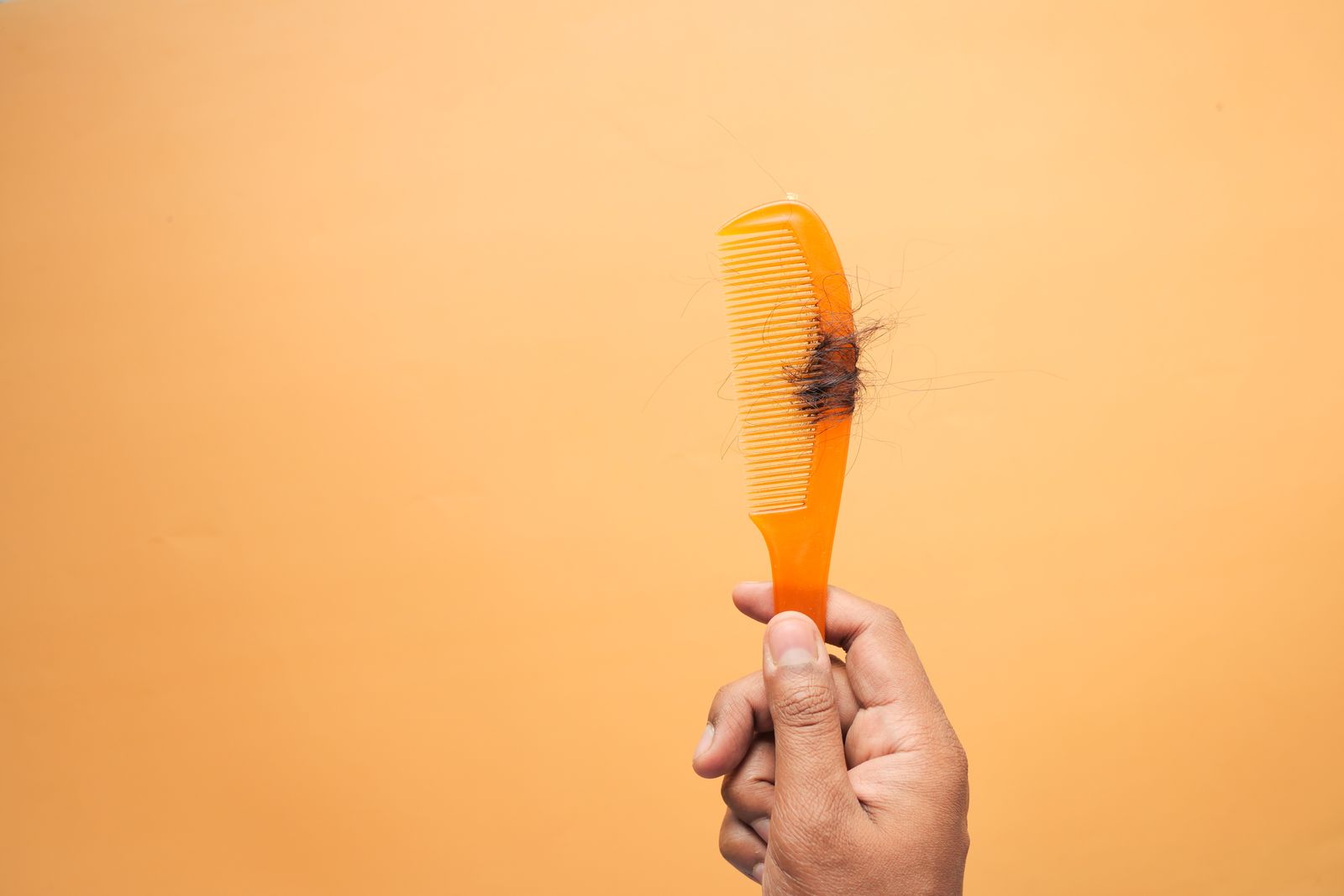 Close-up of a hand holding a plastic comb with loose hair strands caught in the teeth against an orange background