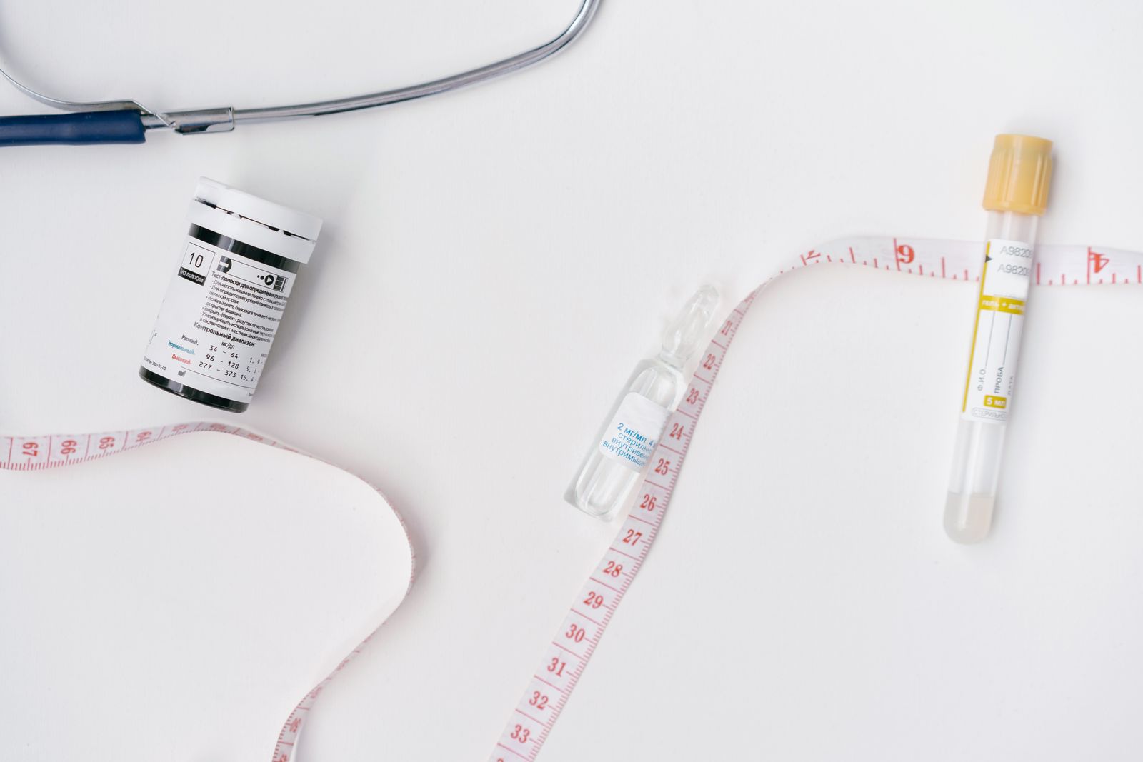 A cloth tape measure looped across a white surface beside a glass medication ampoule, a small sample vial, and a container of test strips