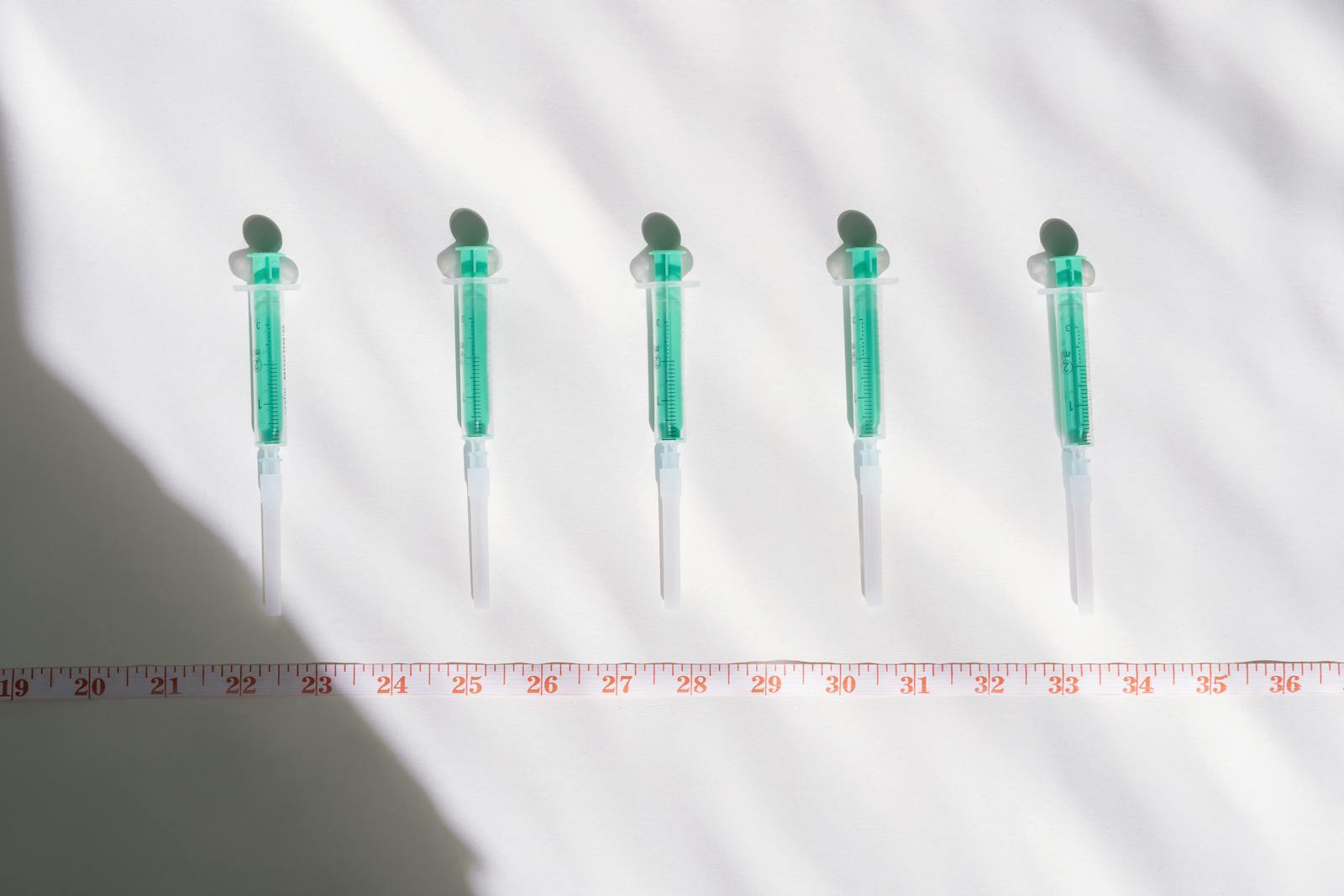 Five sterile syringes arranged in a row above a centimeter measuring tape on a clean white surface, illustrating dosing and measurement precision