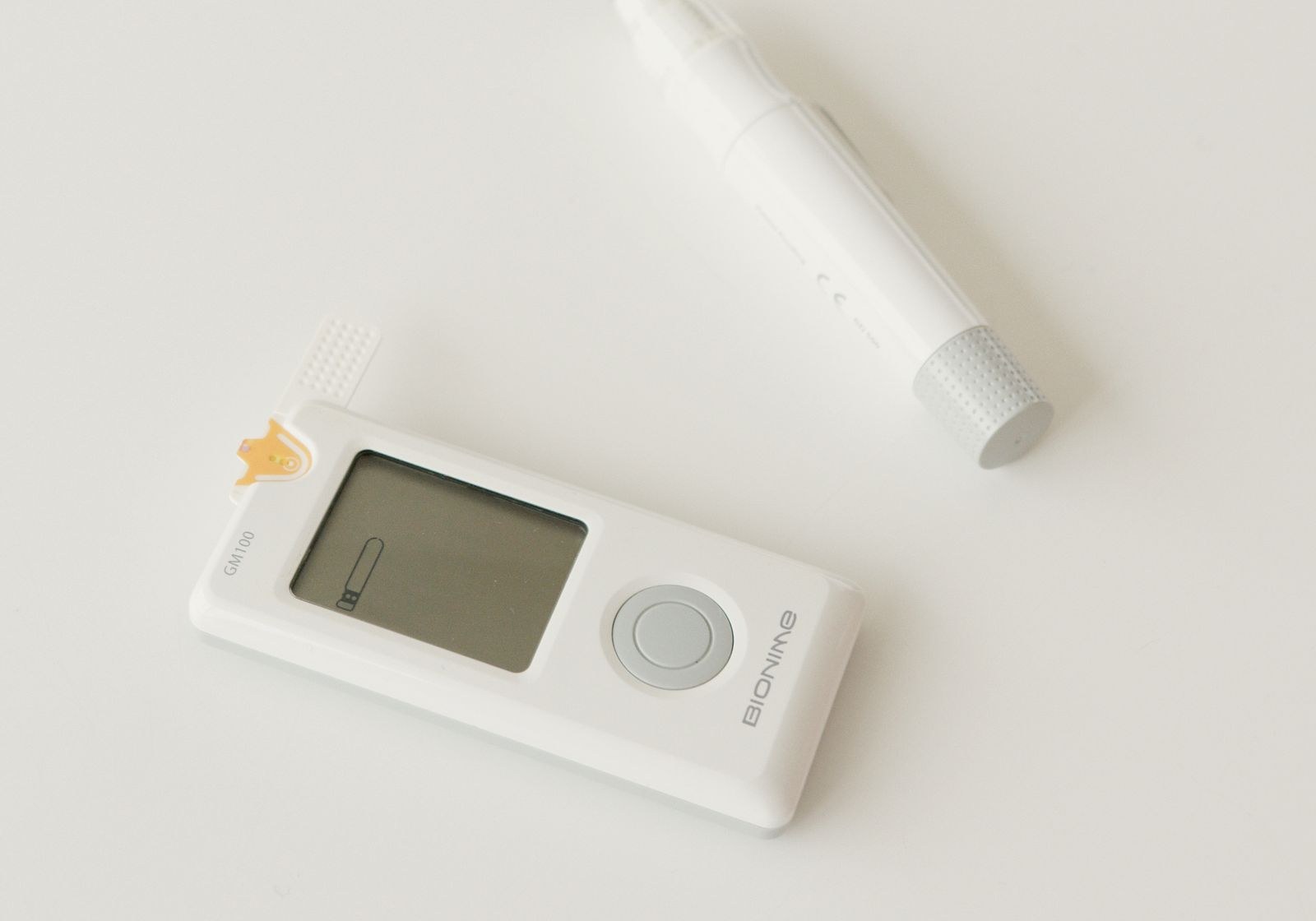 A white handheld blood glucose meter with a test strip inserted, beside a lancing device on a pale surface