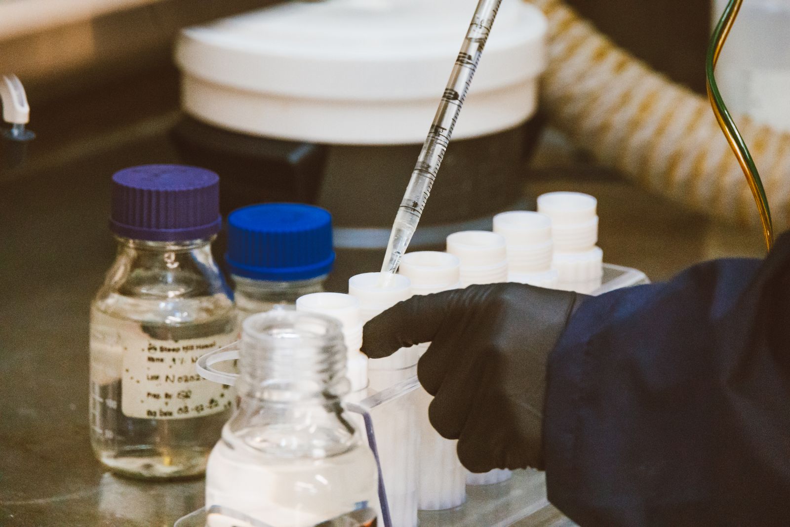 A gloved hand using a pipette to transfer liquid into vials on a lab bench beside reagent bottles, evoking a drug still in research and development