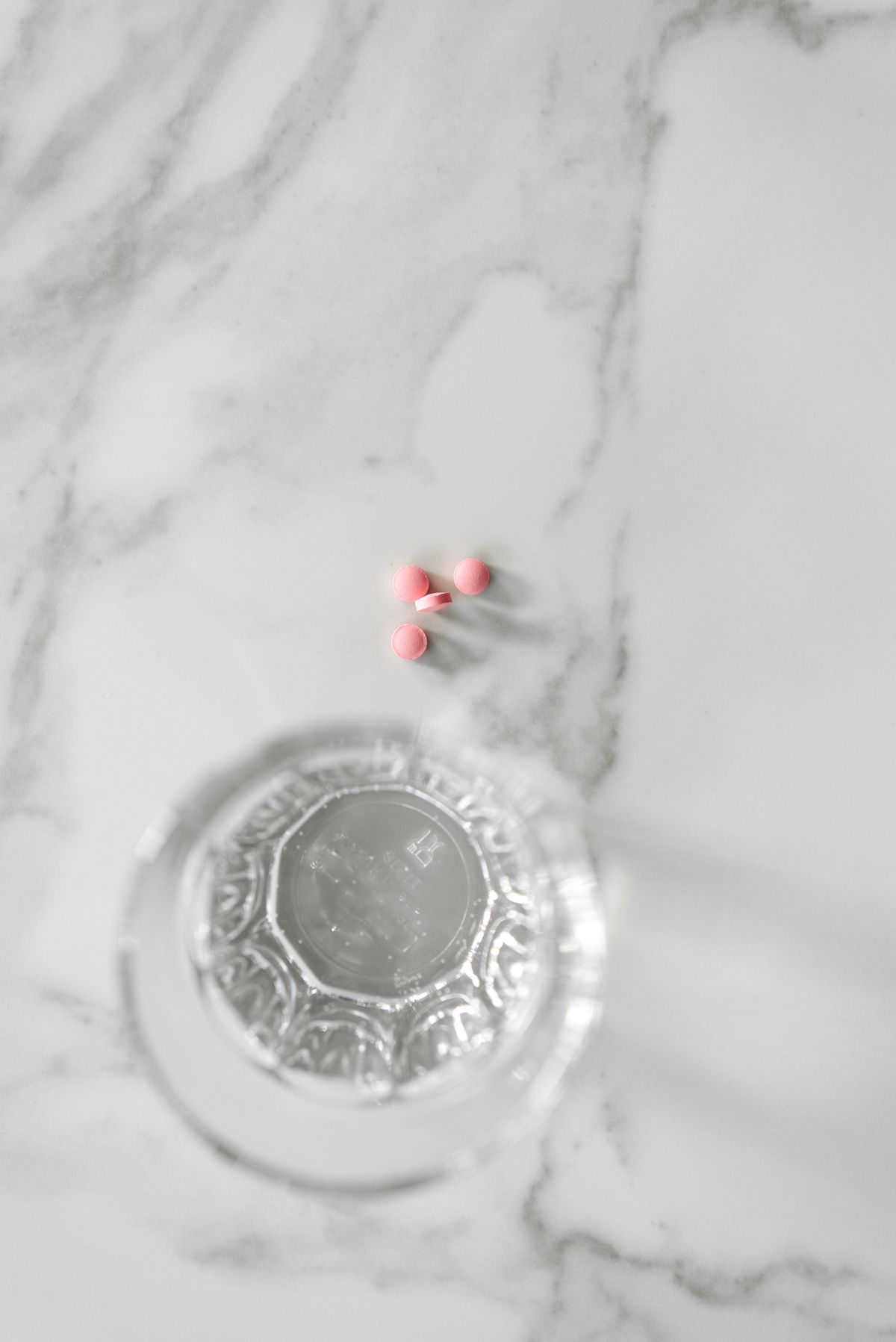 Three small pink tablets beside a clear glass of water on a white marble surface, viewed from above
