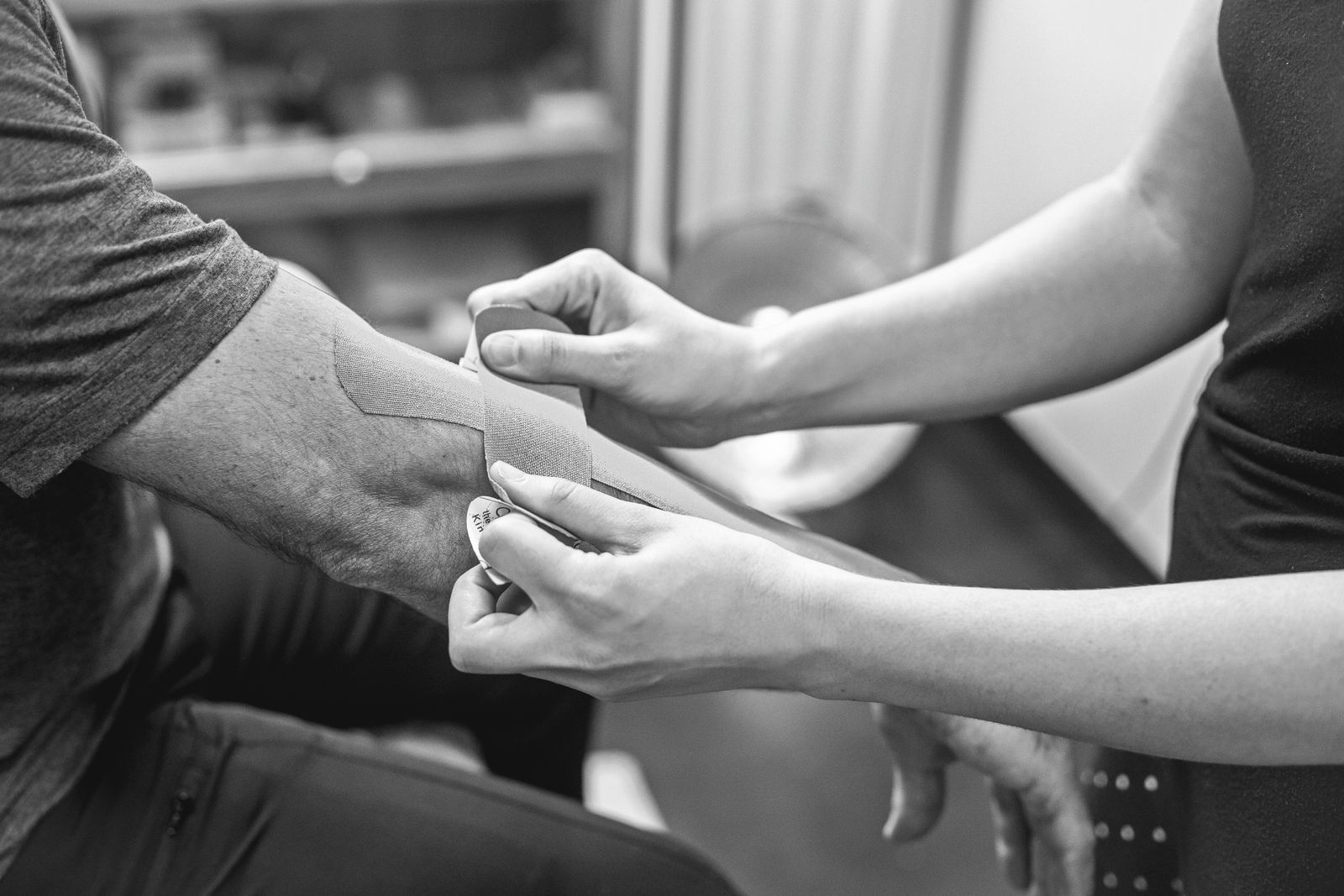 Black-and-white close-up of a therapist's hands applying kinesiology tape to a person's forearm and elbow for tendon support in a clinic