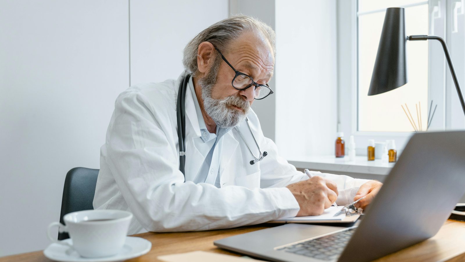 Senior physician at a desk writing a prescription, with a stethoscope and patient chart visible