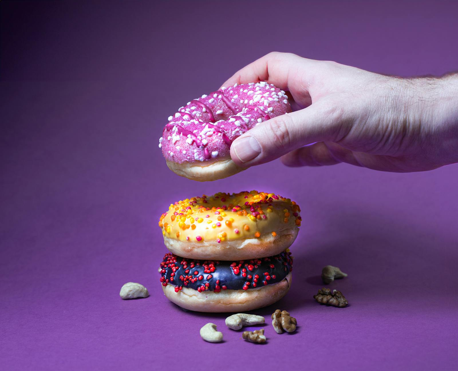 A hand reaching down to pick a pink sprinkled donut from a stack of frosted donuts against a purple background