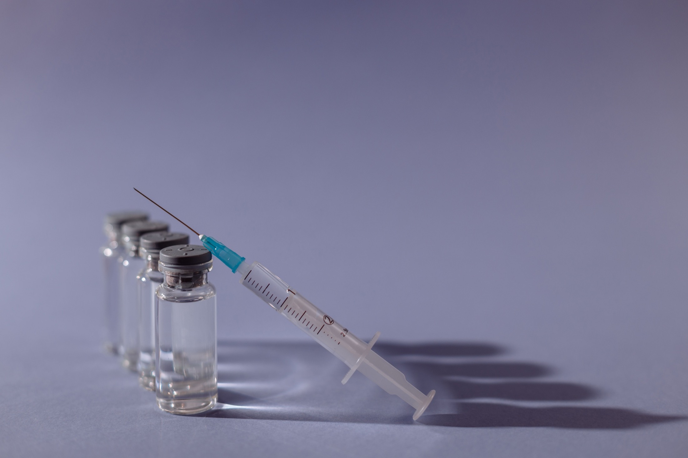 A row of small clear glass vials filled with clear liquid, with a graduated syringe leaning against the front vial on a plain gray surface
